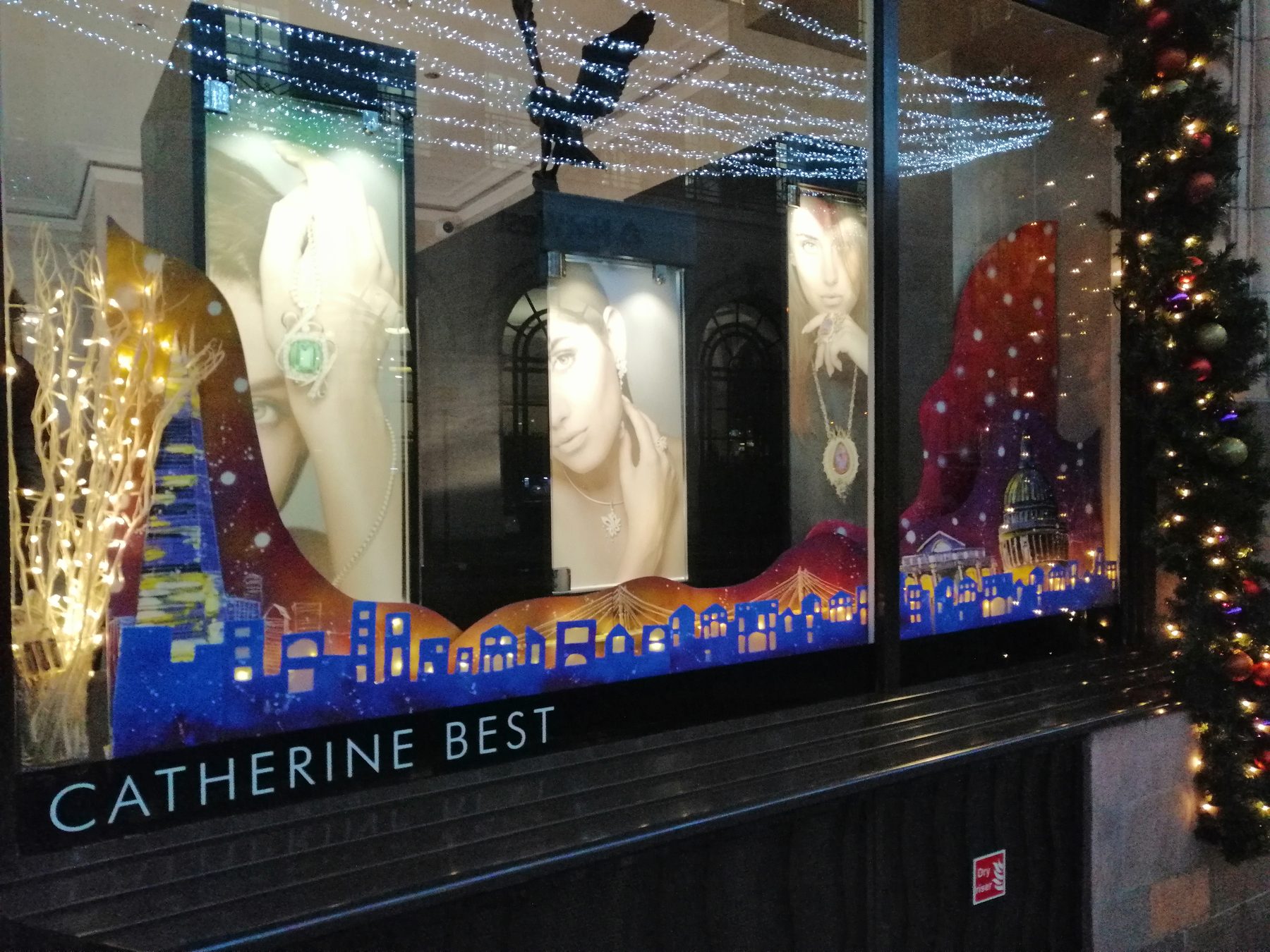 The full Christmas window after dark: a painted London skyline lit from behind